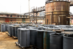 Black barrels containing hazardous waste sitting in front of a large storage container. There are buildings surrounding the area.