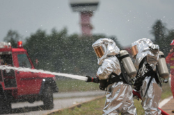 Two firefighters in silver fully-encapsulated suits spray a hazard.