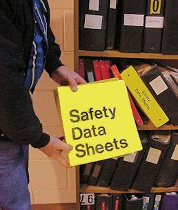 Worker holding yellow binder labeled Safety Data Sheets in front of shelves full of binders