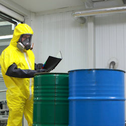 Worker wearing head to toe PPE standing in front of two sealed drums reading paperwork