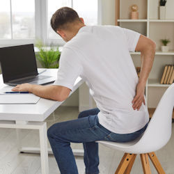 worker sitting at desk in front of computer with hand touching back pain