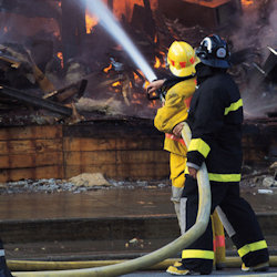 Two firefighters holding hose that is spraying large building fire.