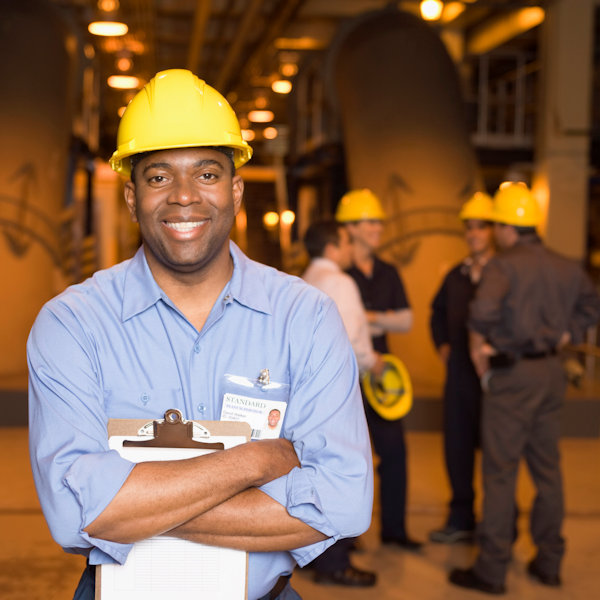 Manager facing forward wearing hard hat holding a clip board. Other employees in the background.