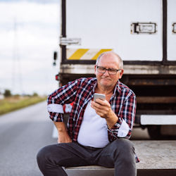 Truck driver sitting on back of truck that is broken down on road.