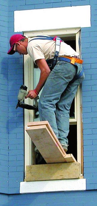 Worker in a very dangerous position standing on a board sticking out a window, holding a nailgun, and wearing a fall harness.