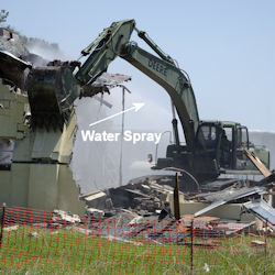 Excavator demolishes building using water to keep dust down.