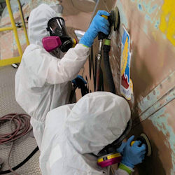 Two workers in full PPE removing paint from wall