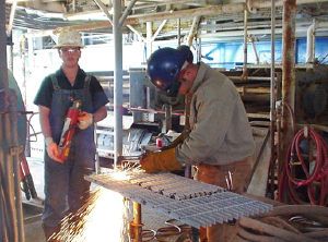 One worker welding and another worker standing by with a fire extinguisher.